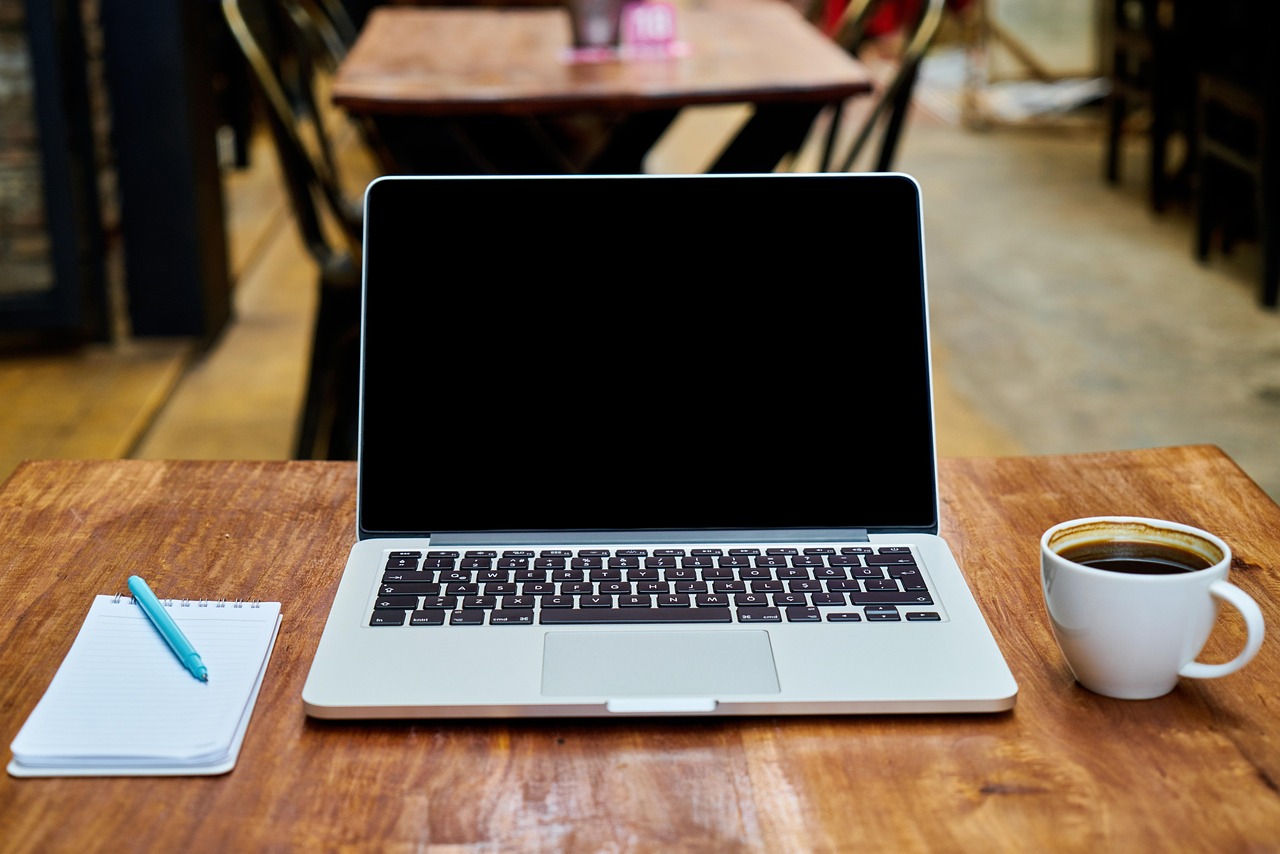 Laptop, üres jegyzettömb, toll és kávéscsésze egy faasztalon.