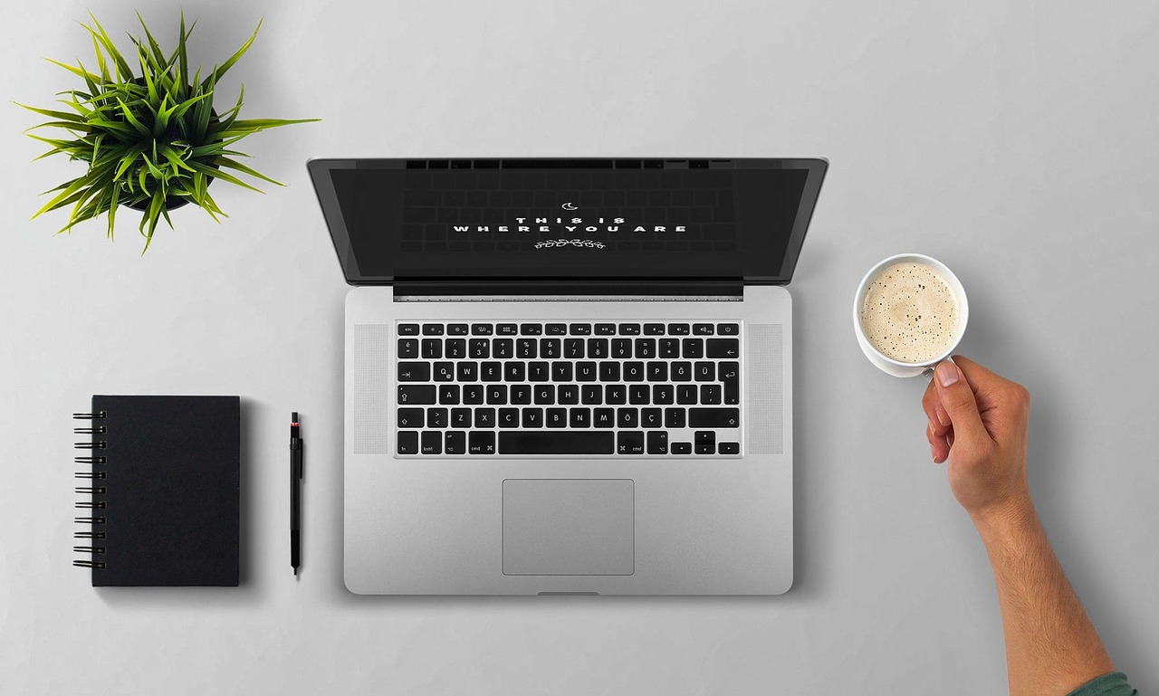 Laptop, jegyzetfüzet és kézben tartott kávéscsésze világos íróasztalon.